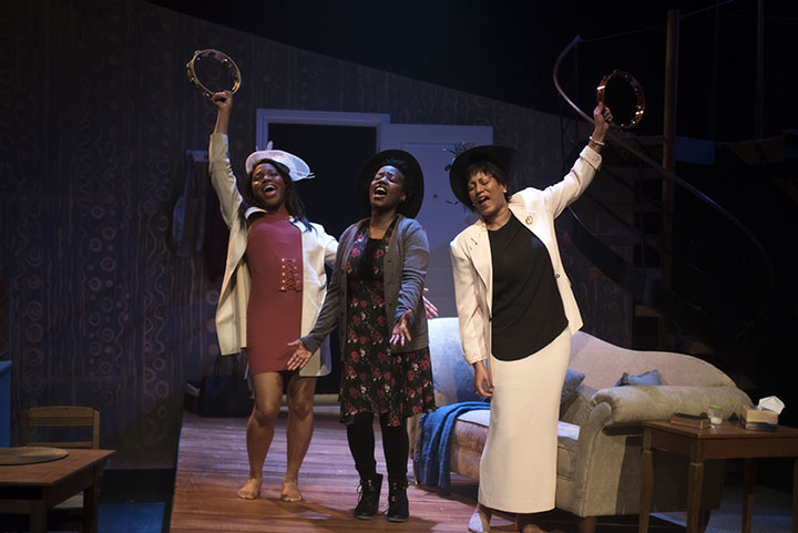 Three black women wearing hats hold their arms in the air and shake tambourines